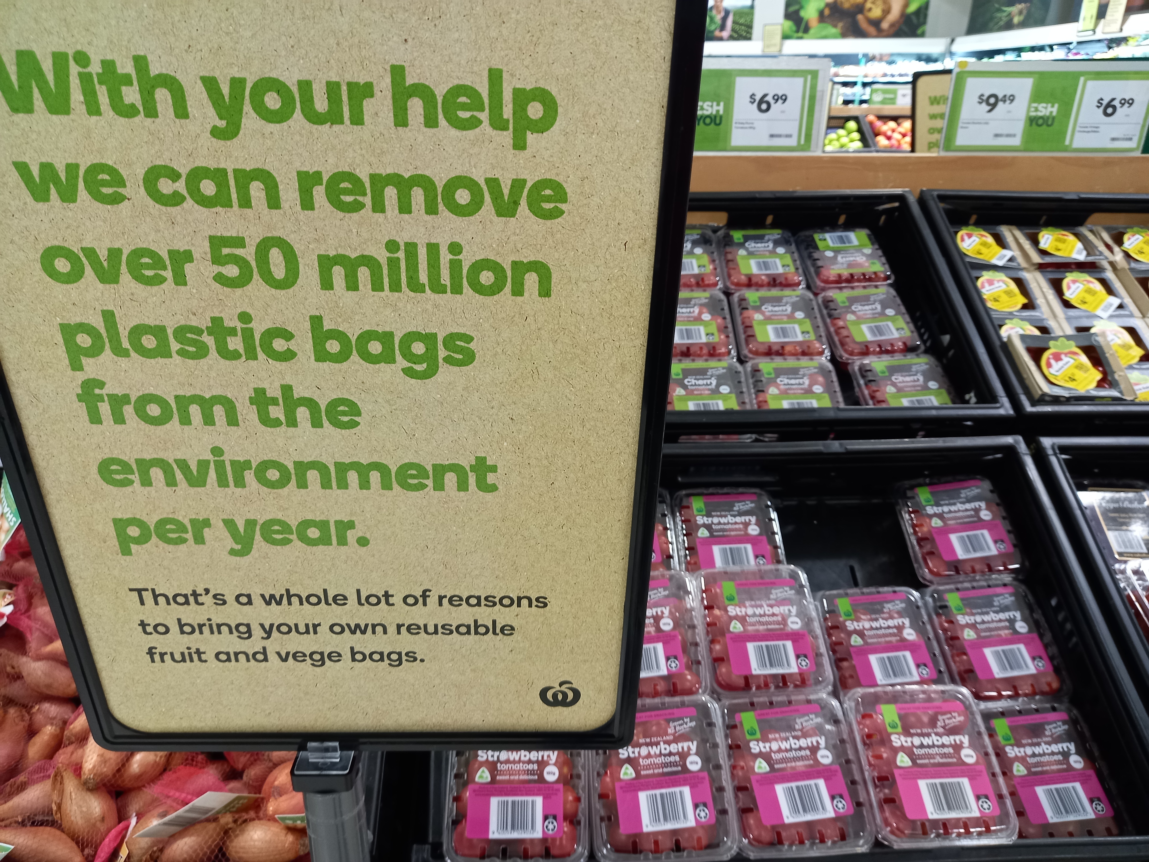 Image: a Coyntdown sign next to a bin of Countdown branded tomatoes in plastic punnets. The sign says "With your help we can remove over 50 million platic bags from the environment each year. Thats a whole lot fo reasons to bring your own reusable fruit and vege bags."